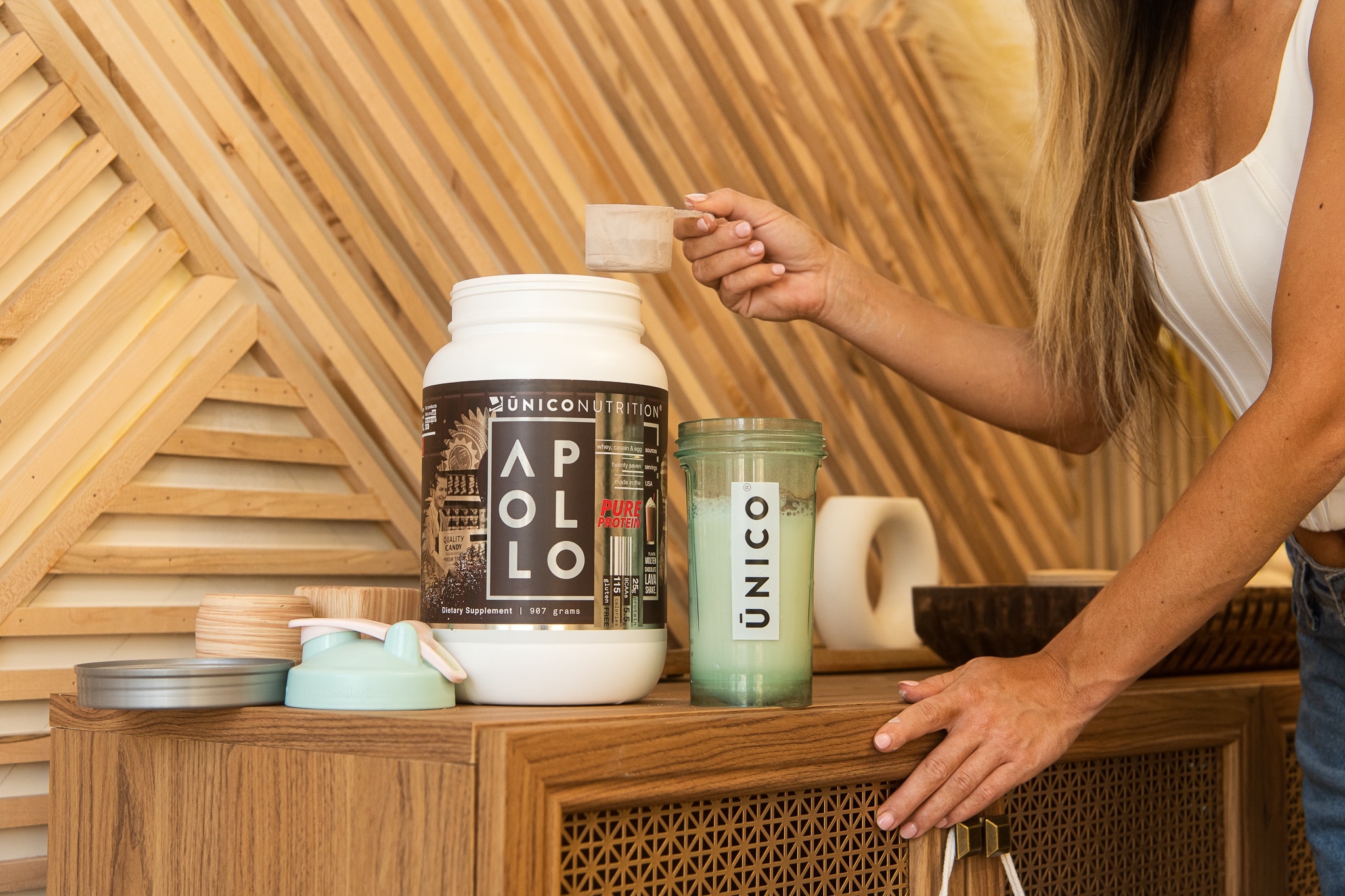 woman scooping protein powder out of a jar into a blue shaker bottle in front of artistic wooden paneling.